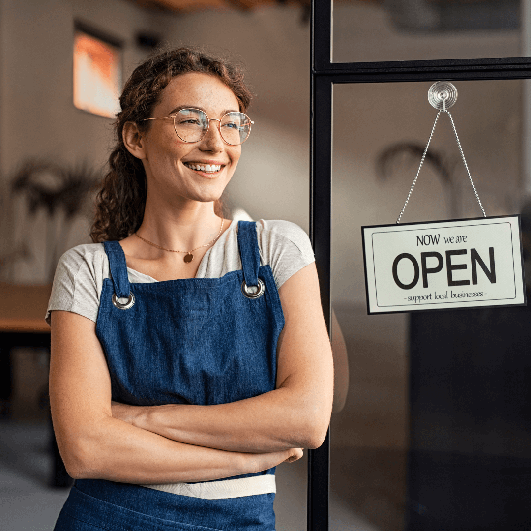 Smiling female small business owner standing proudly by her shop's 'Open' sign, illustrating Hebden Legal's guidance on commercial leases and business structuring in Dunsborough and the wider South West WA region.